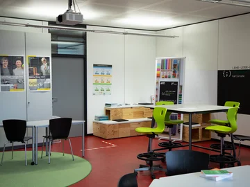 Ein Seminarraum mit flexibel angeordneten Tischgruppen, einem Flipchart und Plakaten an den Wänden. 