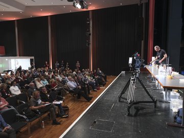 Ein Vortragender steht auf dem Podium, Ein Vortragender steht auf dem Podium, vor ihm stehen chemische Laborgeräte, rechts ist das Publikum zu sehen.