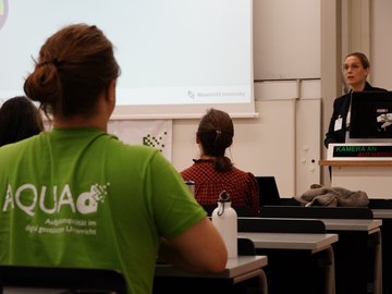 Links im Vordergrund des Bildes sitzt eine Person mit grünem T-Shirt, auf dem auf der Rückseite "AQUA-d" steht. Er schaut nach vorne, wo eine Person an einem Pult steht und einen Vortrag hält.