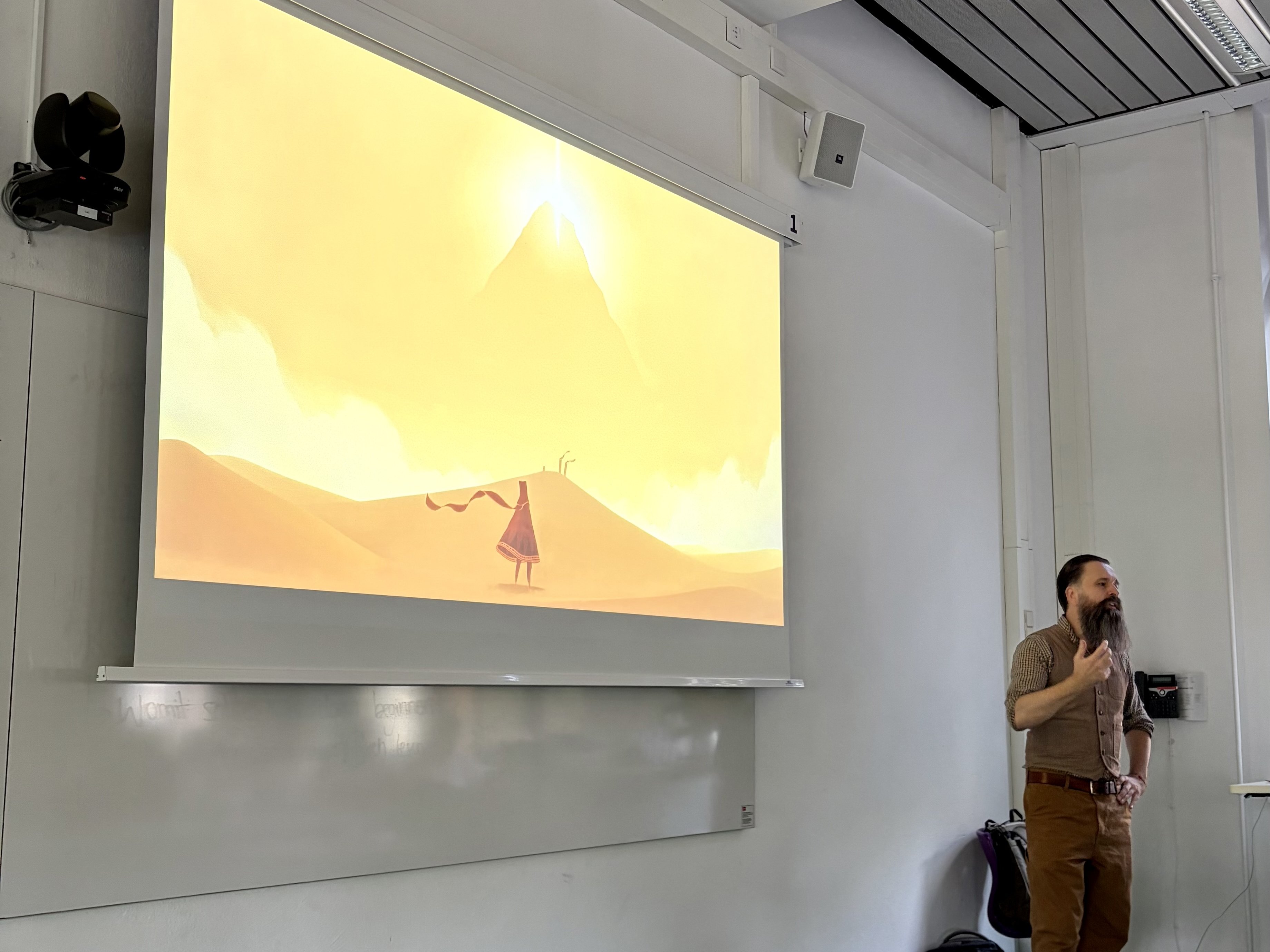 Jörg Luibl hält einen Vortrag, auf dem Beamer neben ihm ist ein Videospiel zu sehen