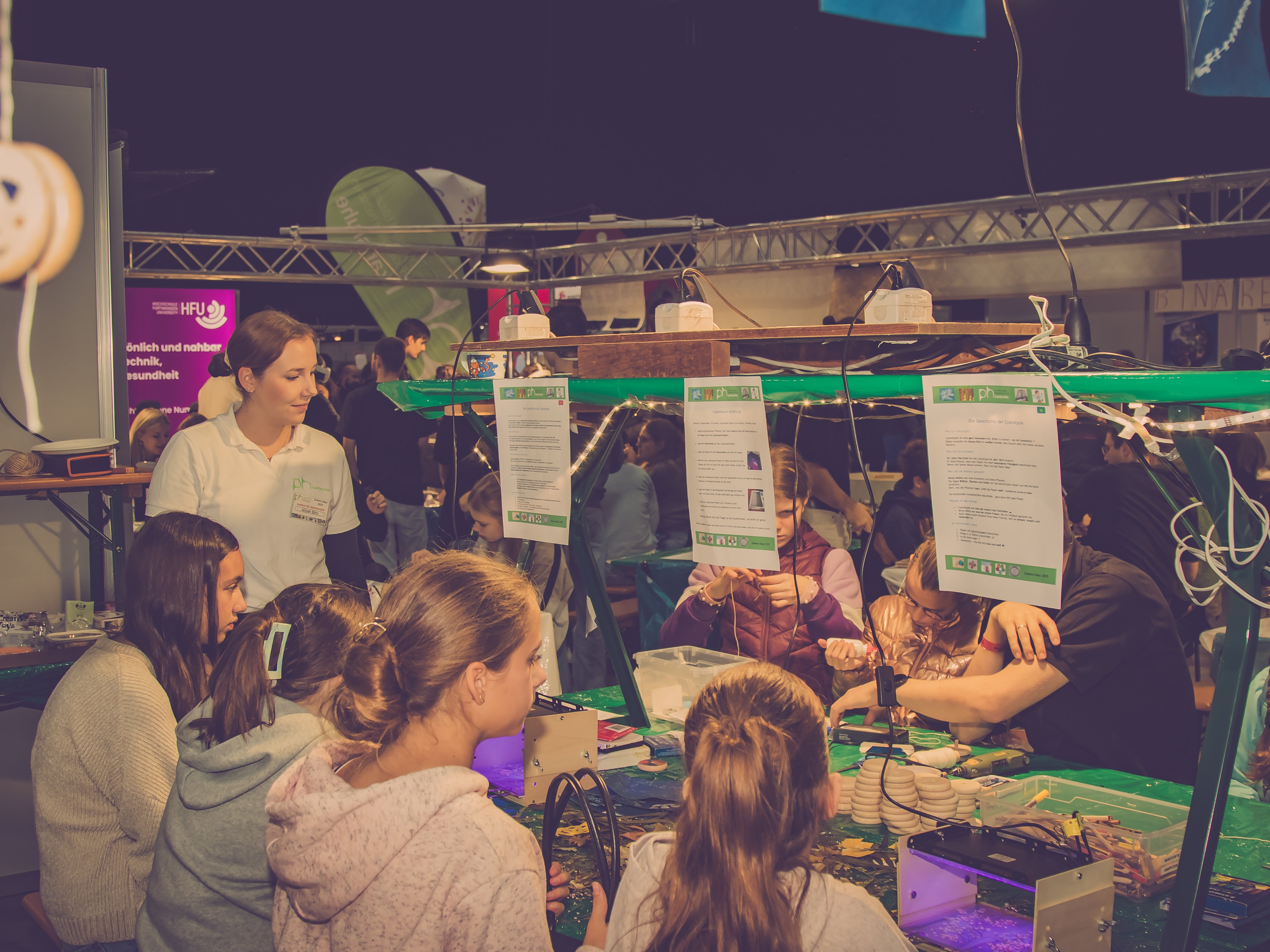 In einer großen Halle sind an einem Tisch mit mathematischen Spielereien sitzend zu sehen, eine Studentin steht rechts und erklärt die Aktivität.