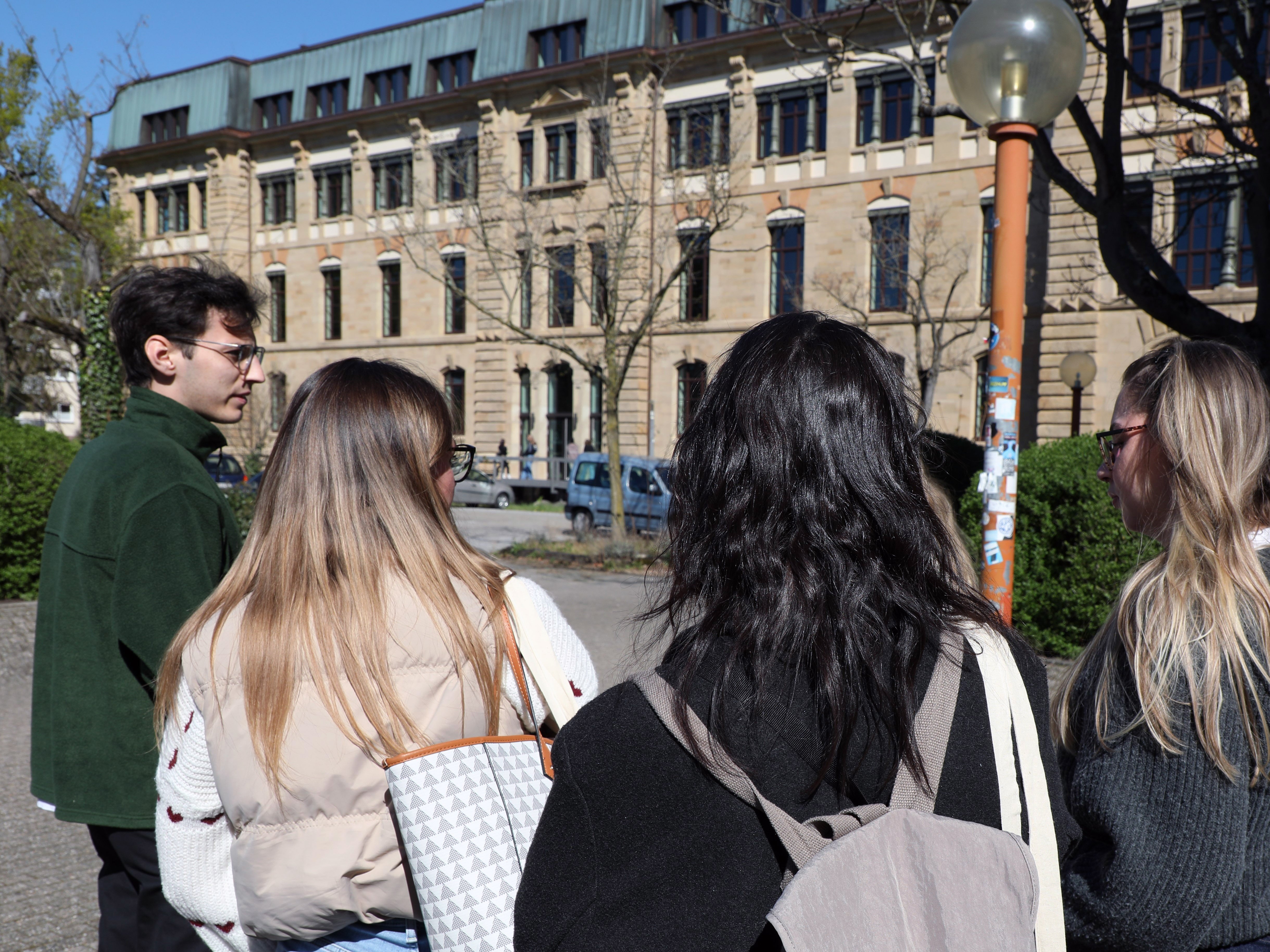 4 Studierende, 3 junge Frauen mit langen Haaren und ganz links ein junger Mann, sind von hinten zu sehen, wie sie auf das Hauptgebäude der PHKA zugehen.