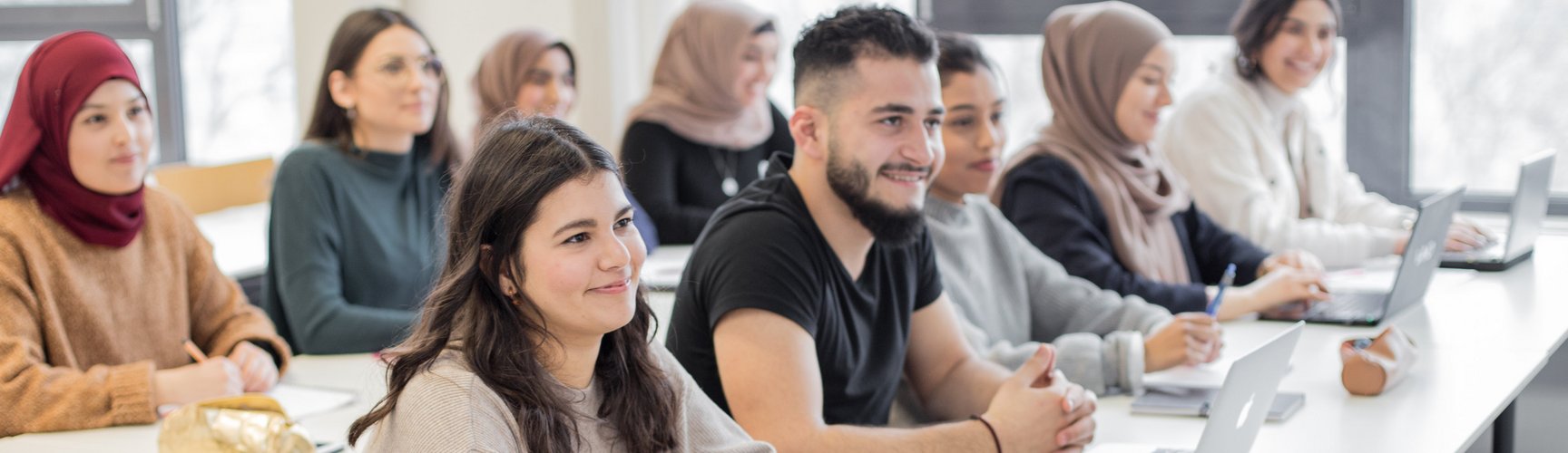 Studierende sitzen in zwei Tischreihen in einem Seminarraum, auf den Tischen Laptops und Schreibblöcke.