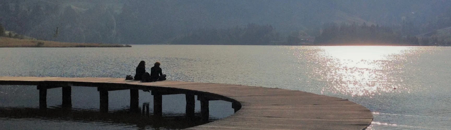 Ein großer gebogener Steg an einem See, in der Mitte sitzen zwei Menschen, im Hintergrund mittelgroße Berge.