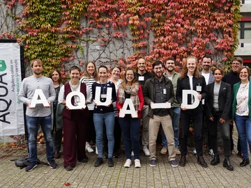 Die Promovierenden und Leitungspersonen des FuN-Kollegs "AQUA-d" stehen vor einer herbstlich bewachsenen Wand im Freien. Links steht eine Beachflag mit dem Logo des Kollegs. Die Personen in der ersten Reihe halten weiße Holzbuchstaben, die "AQUA-D" buchstabieren.  