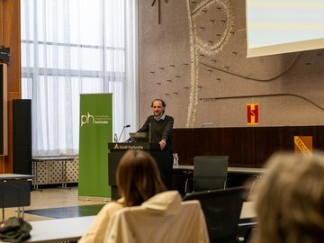 Vortrag von Prof. Bernhard Standl. Er steht am Rednerpult, links ein RollUp mit PH Logo.