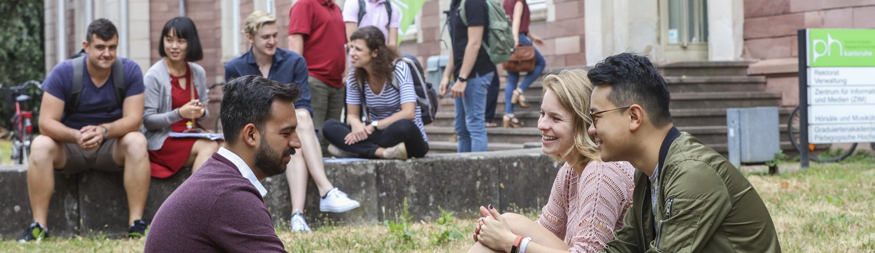3 Studierende sitzen auf der Wiese vor dem Haupteingang der PHKA