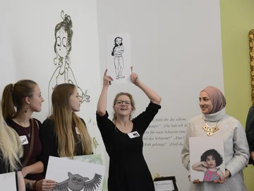 Eine junge Frau hält eine Zeichnung von einer Frau im Comic-Stil in die Höhe und erklärt etwas, drei weitere Frauen hören ihr zu.