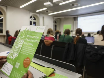 Jemand hält einen Flyer mit dem Titel "Migration, Demokratie, Bildung", im Hintergrund sitzen Studierende in einem Hörsaal.