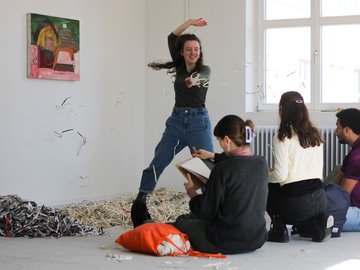 Eine Studentin wirf Papierschnipsel, von denen ein Haufen auf dem Boden liegt; vor ihr sitzen mehrere Studierende.