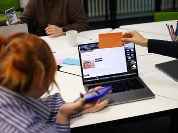 Mehrere Personen sitzen an einem Tisch und arbeiten gemeinsam an einem Laptop, auf dessen Bildschirm eine Anwendung mit Text und einer Zeichnung zu sehen ist. Eine Person hält eine orangefarbene Karte vor den Bildschirm, während eine andere ein Smartphone in der Hand hat.