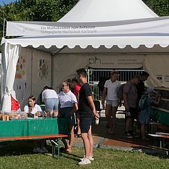 Das Pagodenzelt Mathekosmos im Experimentepark von EFFEKTE. Vordem Zelt ein Biertisch mit einer Kiste mit bunten Bausteinne zum mathematische Muster legen, im Zelt selbst weitere mathematische Spiele, rechts Steinplatten, an denen ein Kind gerade ägyptische oder römische Zahlen hineinmeißelt. Es sind weitere Kinder und Erwachsene im und vor dem Zelt zu sehen.  