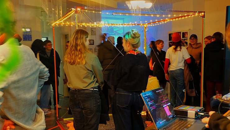 Situation auf der Kunstausstellung mit ein paar Personen, die eine Installation bettrachten, und weiteren im Hintergrund.