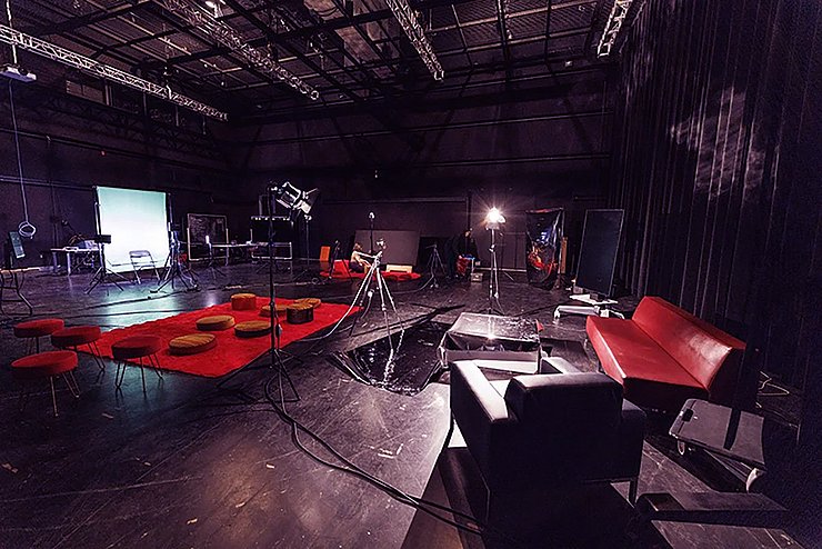 Das Bild zeigt einen großen, dunklen Studio- oder Theaterraum mit hoher Decke. Auf dem Boden stehen verteilt Kamera- und Lichtstative sowie viele Kabel. In der Mitte liegt ein roter Teppich mit runden Sitzkissen und kleinen Hockern. Links hinten ist ein grüner Hintergrund (Greenscreen) mit Stühlen und Technik davor. Rechts im Vordergrund stehen ein rotes Sofa, ein schwarzer Sessel und ein kleiner Tisch. Der Raum wirkt wie eine provisorische Bühne oder ein kreatives Setup ohne Menschen.