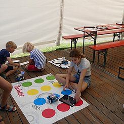 Blick in das Pagodenzelt des Lehr-Lern-Labors Informatik bei EFFEKTE 2023. Im Hintergrund eine Biertischgarnitur mit Tablets und Unterlagen, vorne Kinder platziert um eine Twisterfolie mit mBots und Tablets